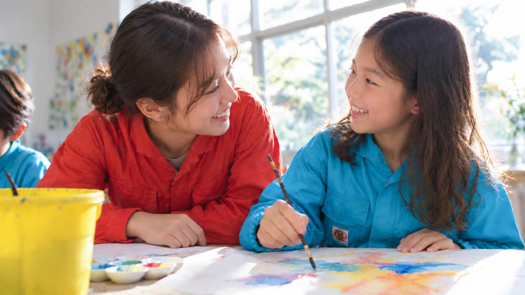 A child painting in watercolor, with a smiling teacher beside them offering a word